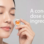 Model with eyes closed holding a single-dose skincare capsule containing a concentrated amber-colored active ingredient.
