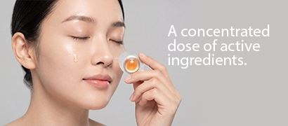 Model with eyes closed holding a single-dose skincare capsule containing a concentrated amber-colored active ingredient.